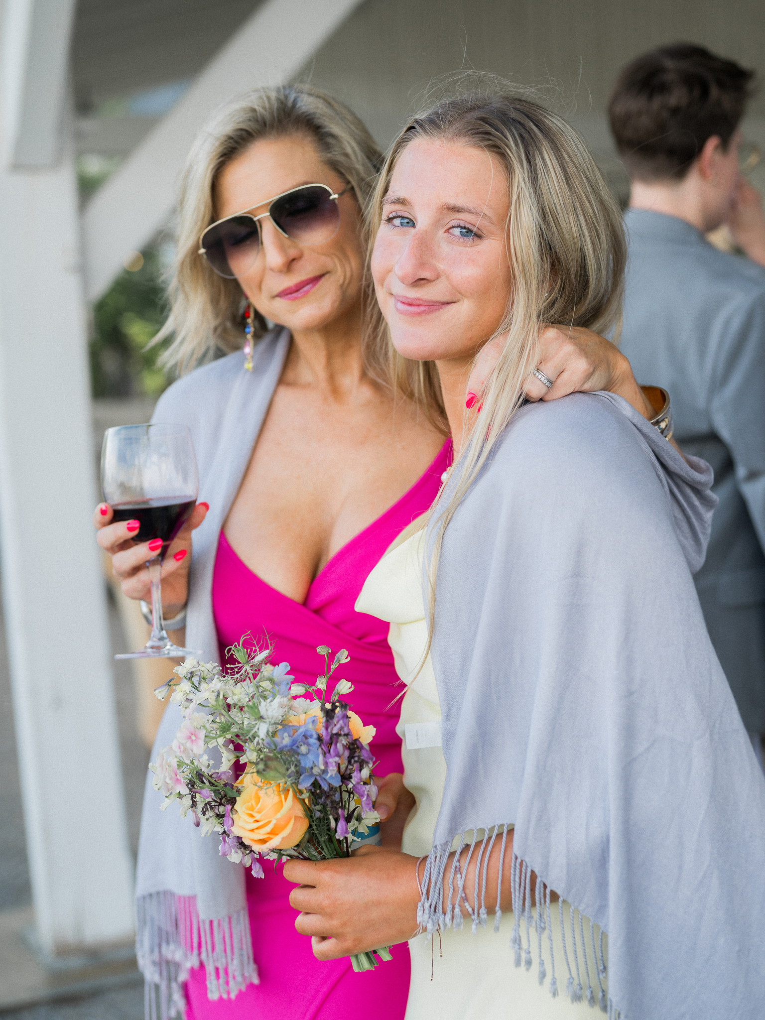 Two wedding guests posing with drinks and a bouquet, dressed in bright colors and smiling during cocktail hour.