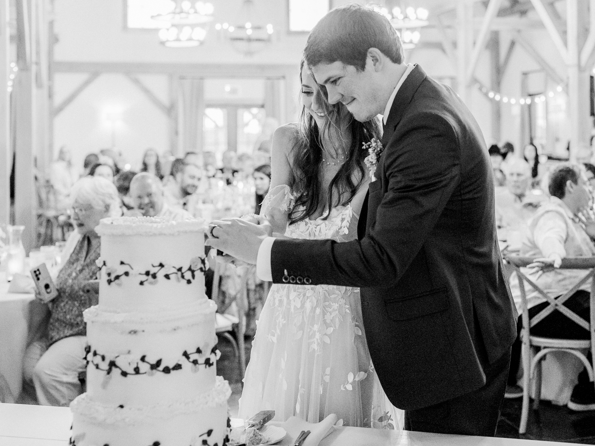 Black and white image of the couple cutting their wedding cake together as guests look on with smiles.