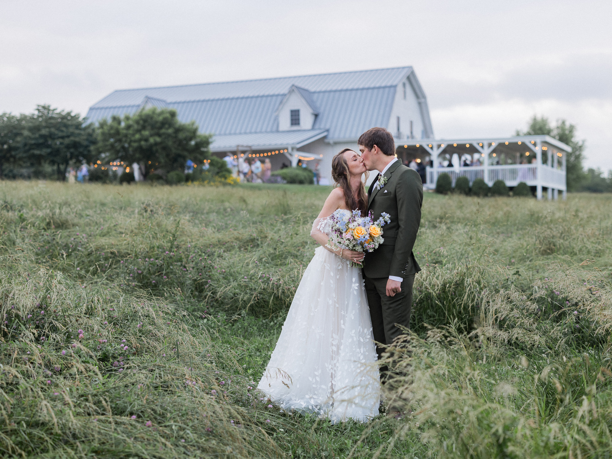 Bride and groom kiss in a grassy field at Blue Bell Farm in front of heir warmly lit reception barn at Blue Bell Farm.
