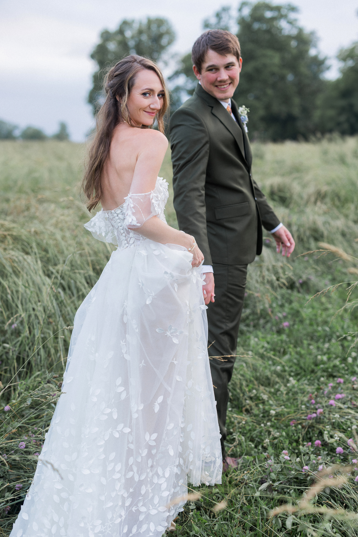 A bride looks over her shoulder back at the camera as she strolls with her groom during golden hour at their Blue Bell Farm wedding.