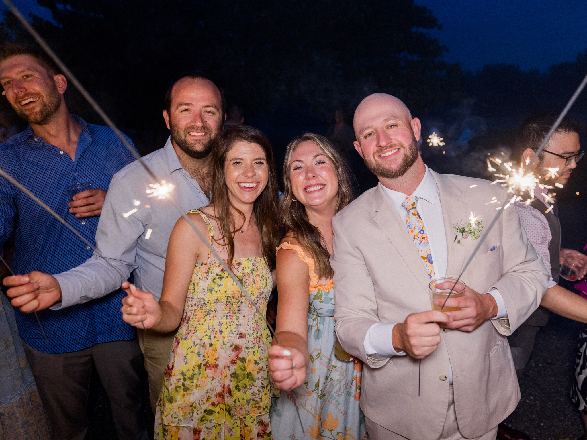 A guest in a cream suit holds a sparkler and a drink, smiling in the middle of a lively sparkler exit at a Blue Bell Farm wedding, documented by Love Tree Studios.