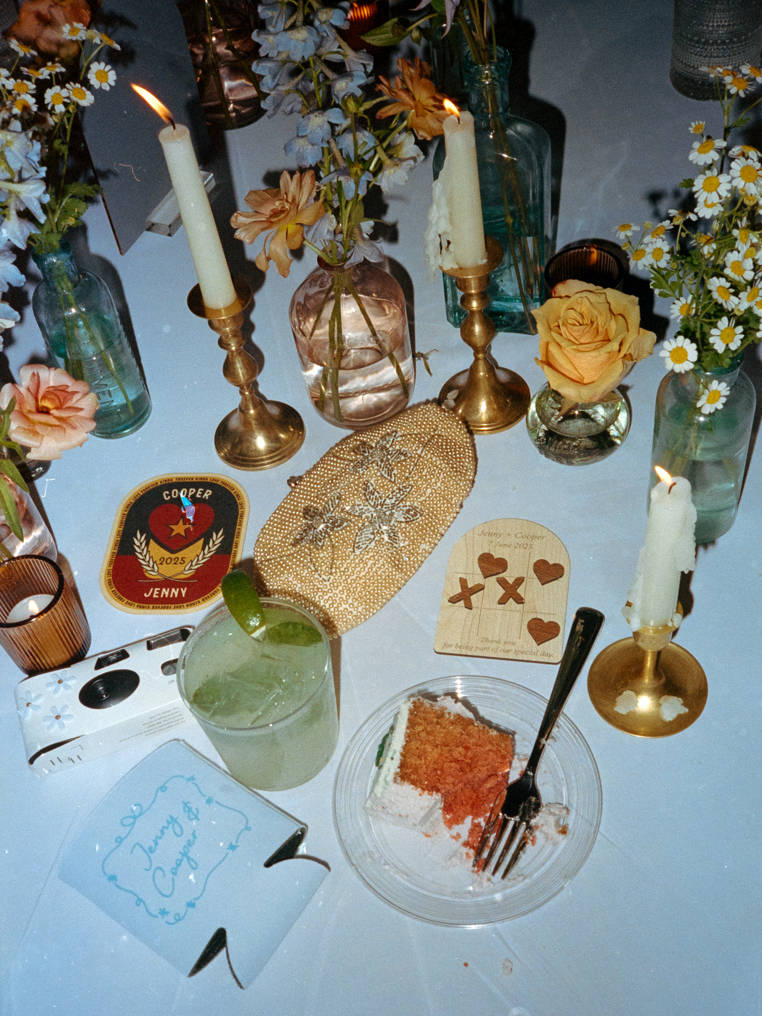 A late night flash flat lay of reception table decor with glowing candles, florals, a cocktail, and a slice of cake during a Blue Bell Farm wedding by Love Tree Studios.