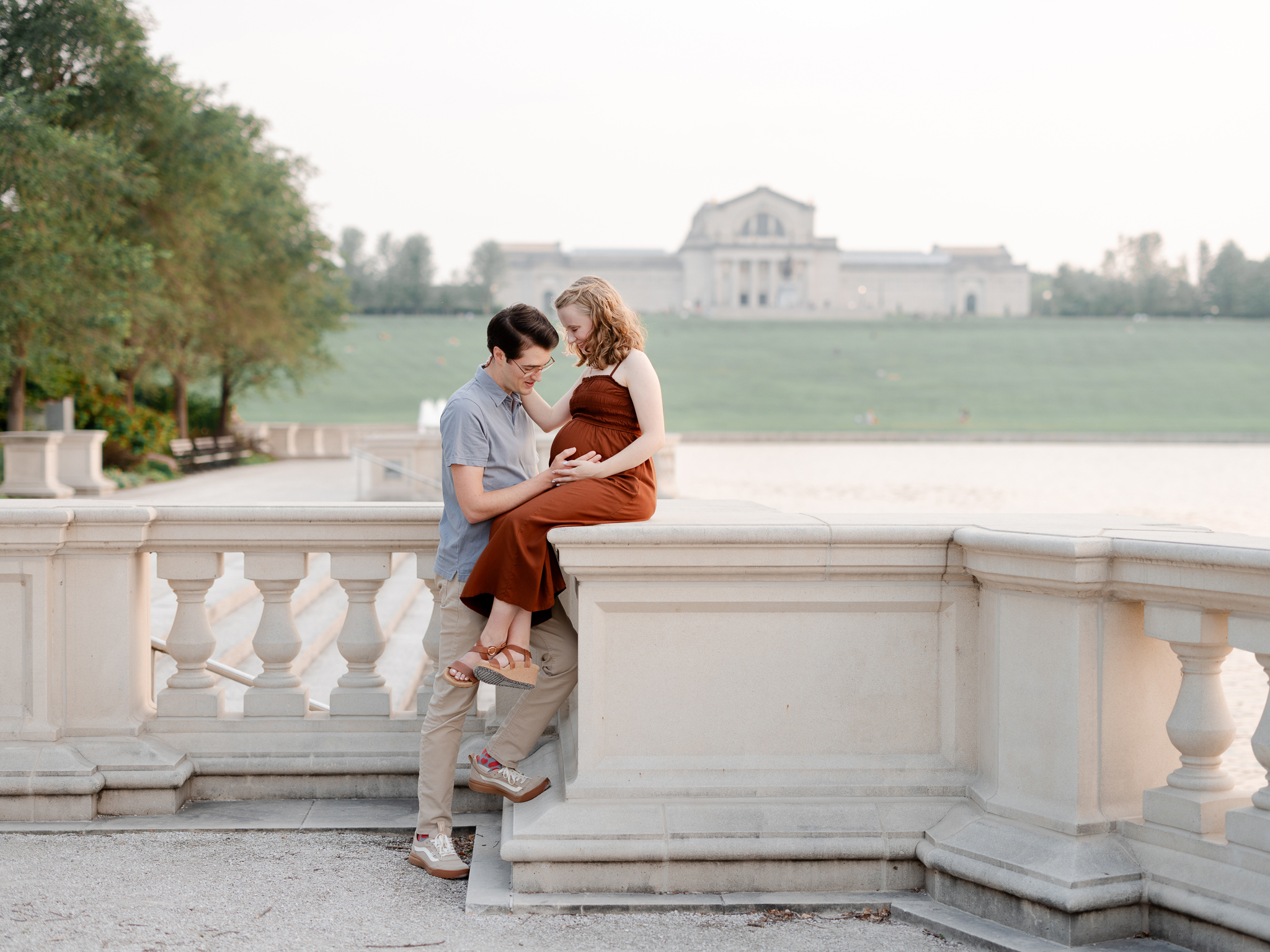 Husband kneels while his wife sits on a stone railing at Art Hill, both holding her baby bump during their maternity photos with Love Tree Studios.