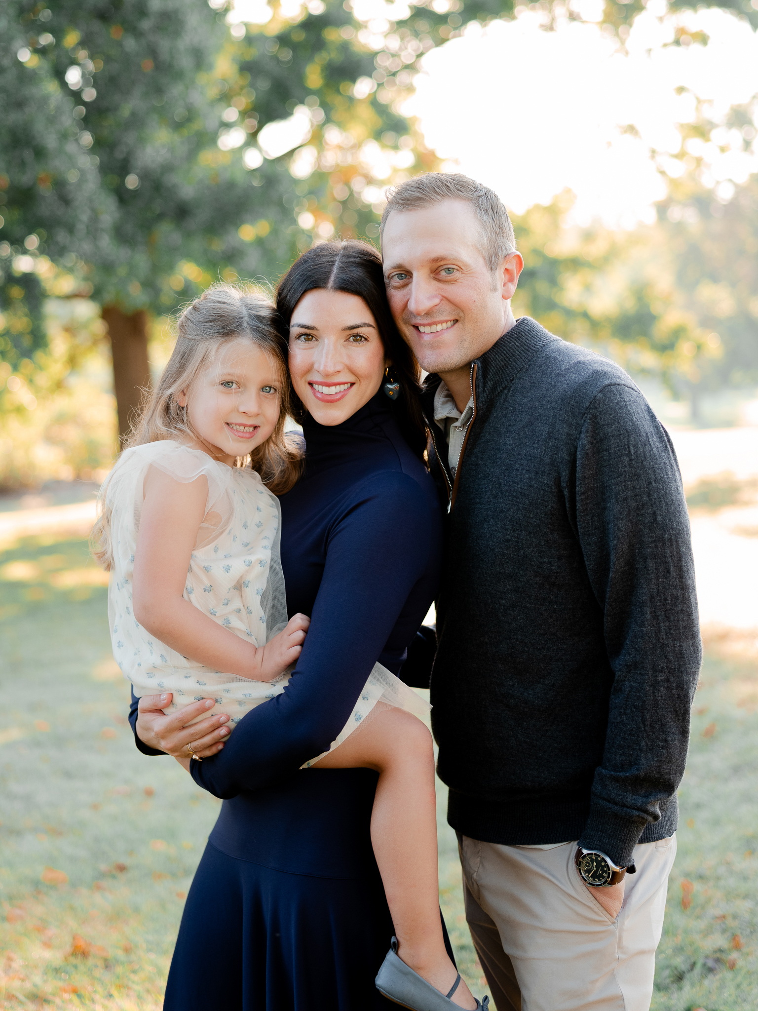 Parents holding their daughter close with trees glowing in morning light – sunrise family photography Columbia Missouri.