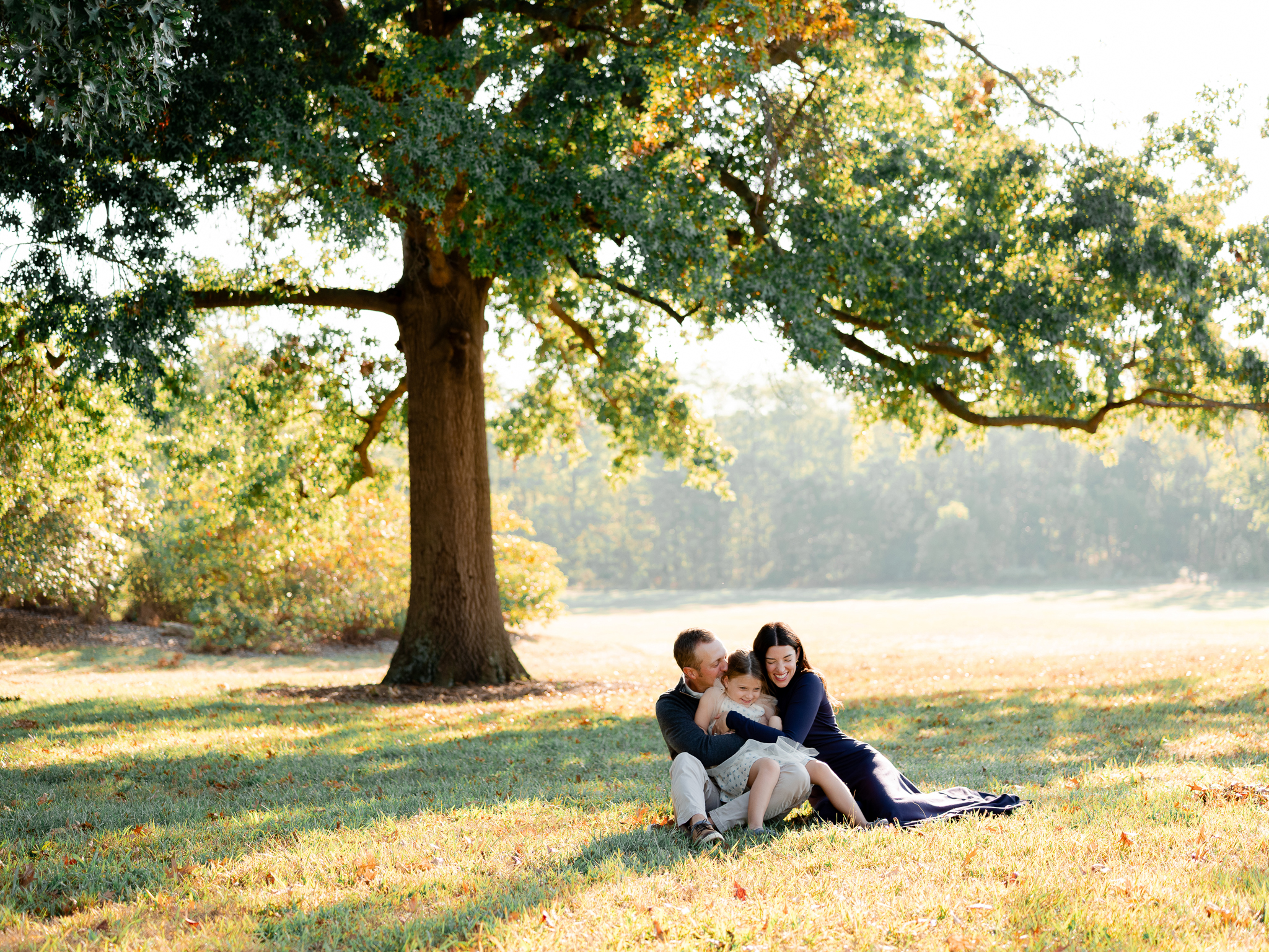 Family sitting together under a large oak tree, cuddling in the morning glow – sunrise family session Columbia Missouri.