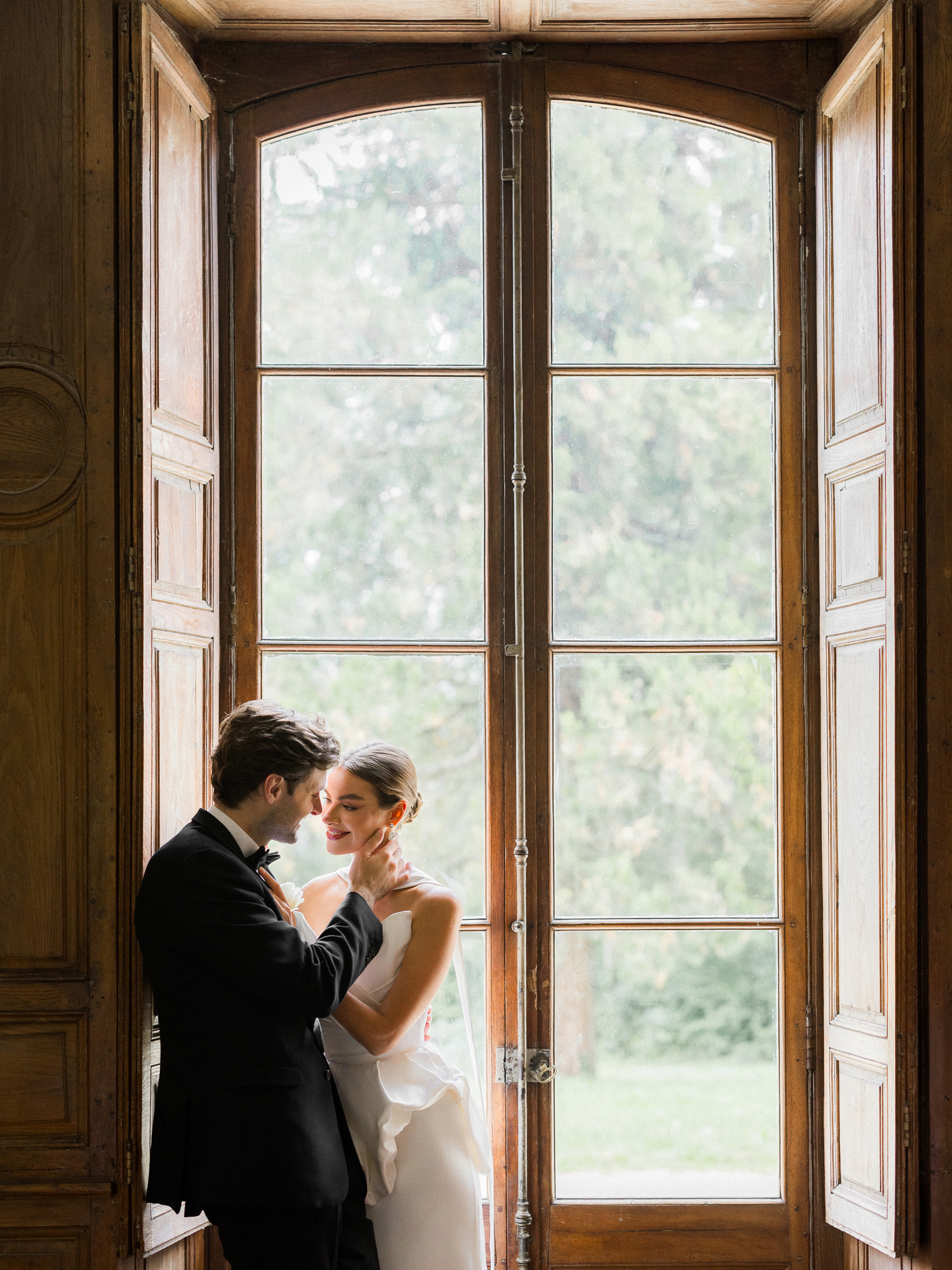 Romantic moment between bride and groom by a tall window at Château de Thauvenay during their Sancerre, France wedding session by Love Tree Studios.