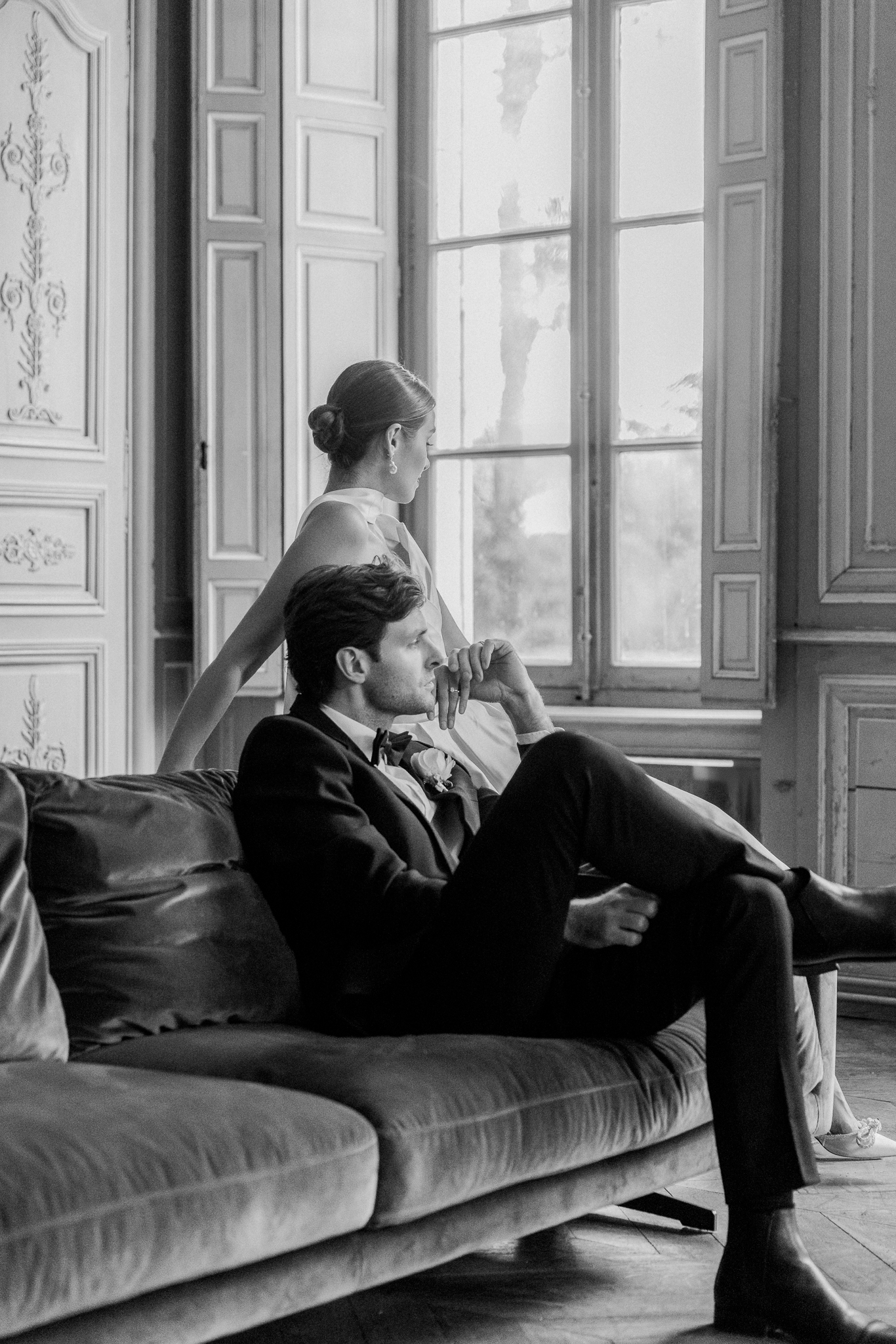Black and white photo of a stylish couple in elegant wedding attire relaxing near tall French windows inside Château de Thauvenay in France.