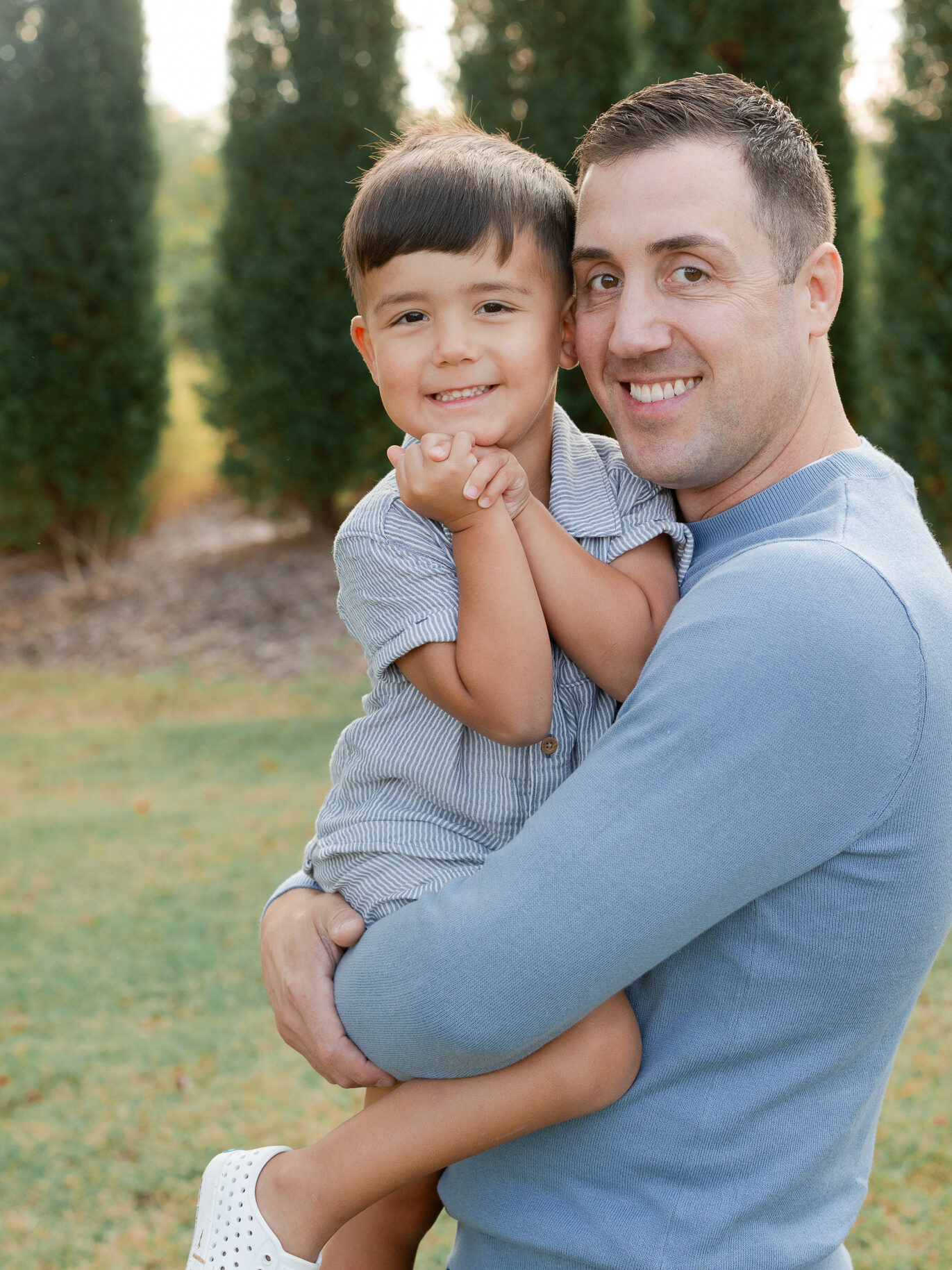 Dad holding son close with tall evergreens behind them during their fall session at Stephens Lake Park.