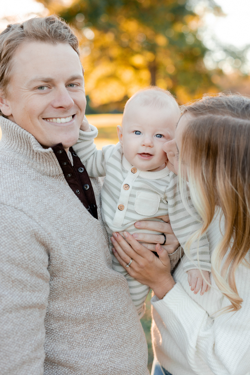 A close up of parents holding their baby as he looks toward the camera while his mother kisses his cheek, taken by Love Tree Studios for golden sunset family photos in Columbia, MO.