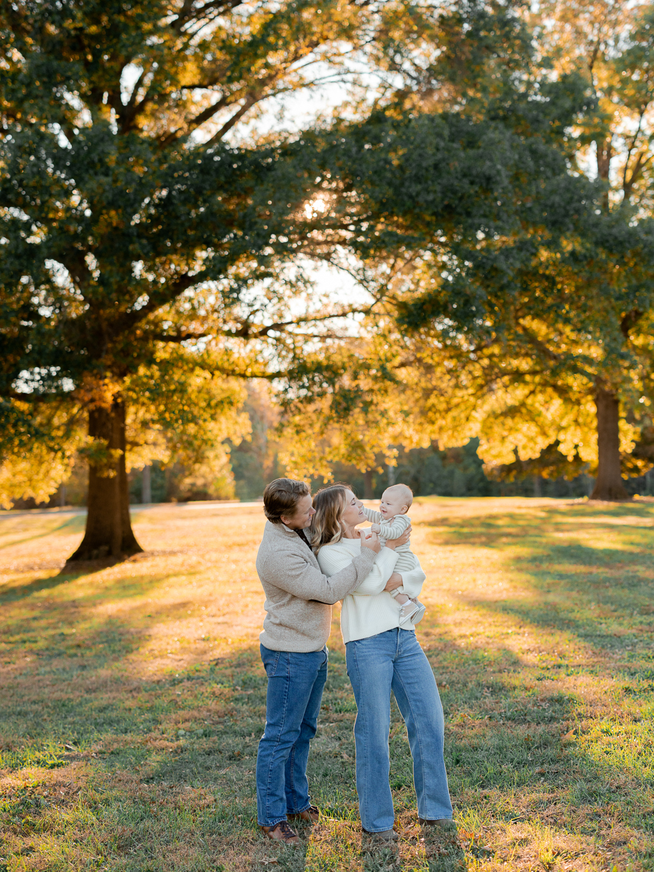 A tender family moment with parents lifting their baby between them under golden fall trees, photographed by Love Tree Studios for golden sunset family photos in Columbia, MO.