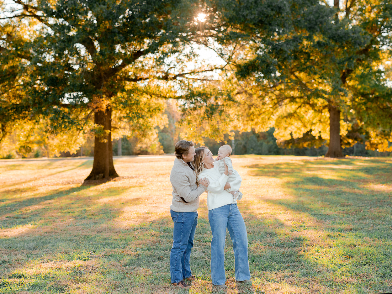 A family standing together beneath glowing fall trees during golden hour, smiling at one another while holding their baby, captured by Love Tree Studios for golden sunset family photos in Columbia, MO.