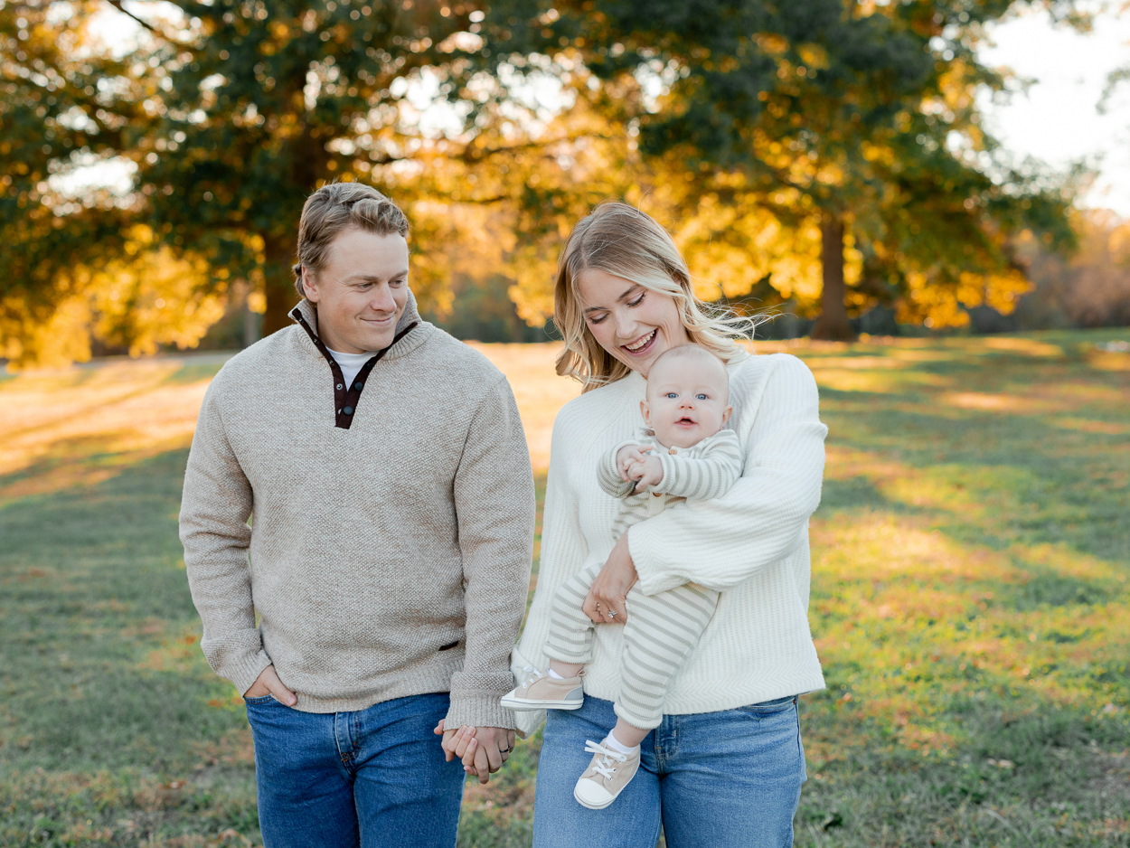 Parents walking through a sunlit field while mom holds their smiling baby, captured by Love Tree Studios for golden sunset family photos in Columbia, MO.