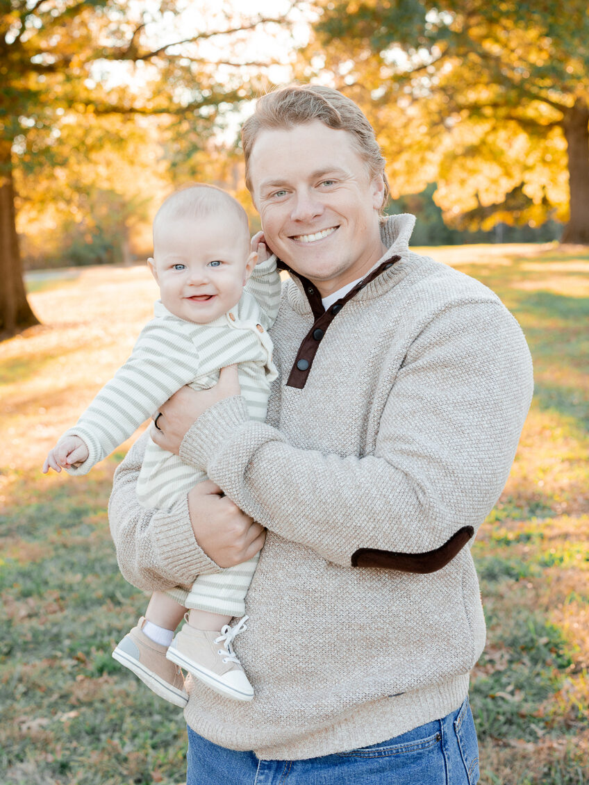 A father holding his baby in a sunlit field with golden trees behind them, captured by Love Tree Studios for golden sunset family photos in Columbia, MO.