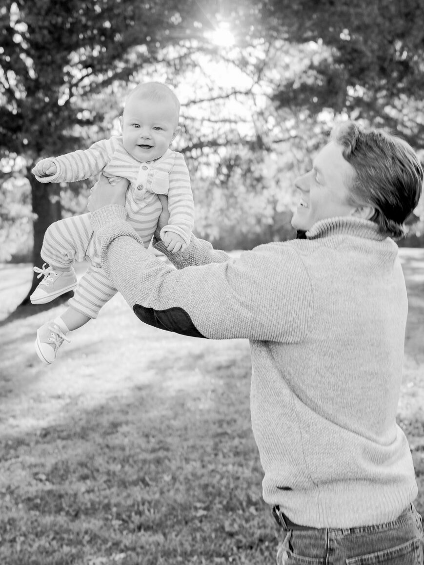 A black and white photo of a father lifting his smiling baby into the air under bright afternoon light, photographed by Love Tree Studios for golden sunset family photos in Columbia, MO.