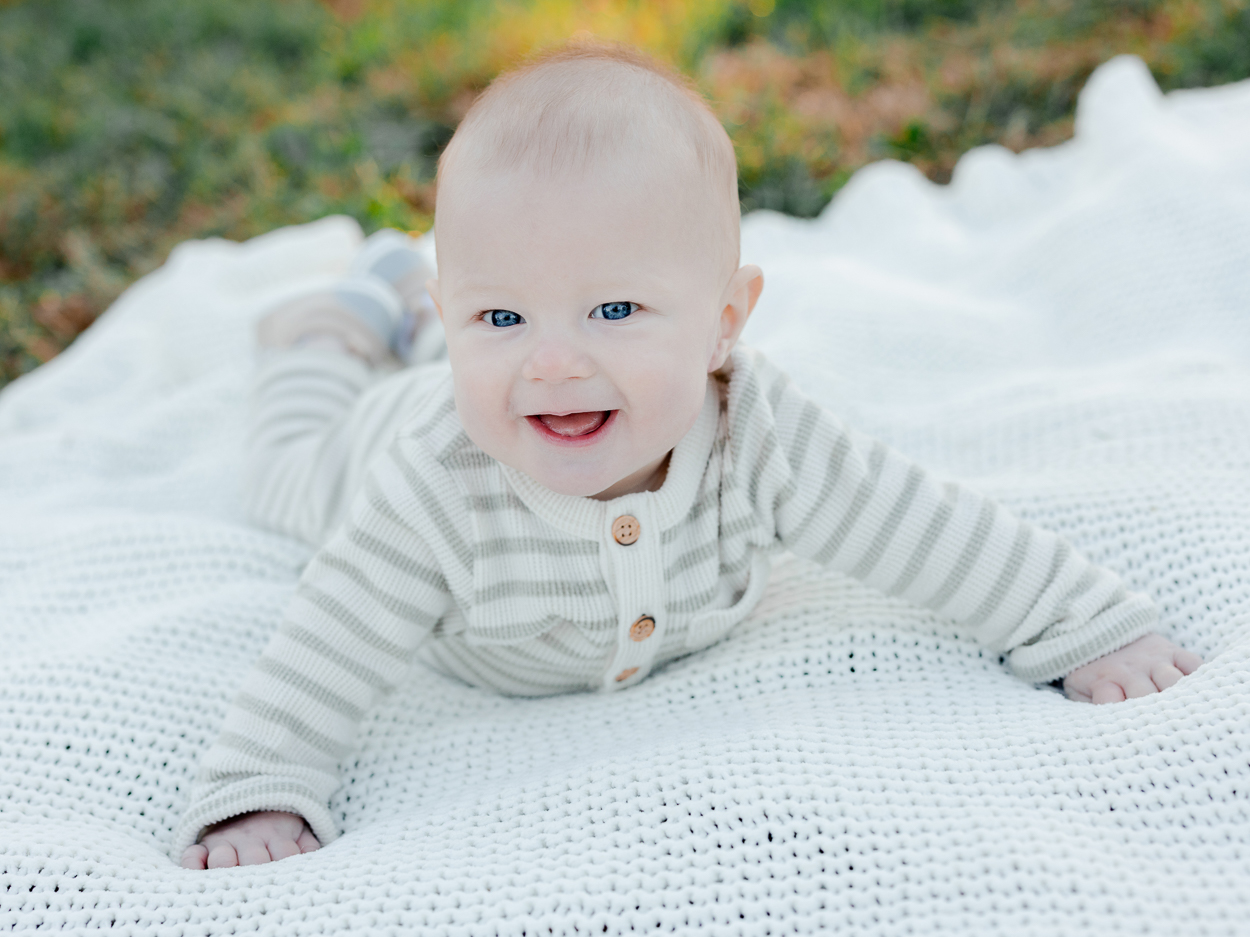 A smiling baby lying on a white blanket in the grass during golden hour, photographed by Love Tree Studios for golden sunset family photos in Columbia, MO.