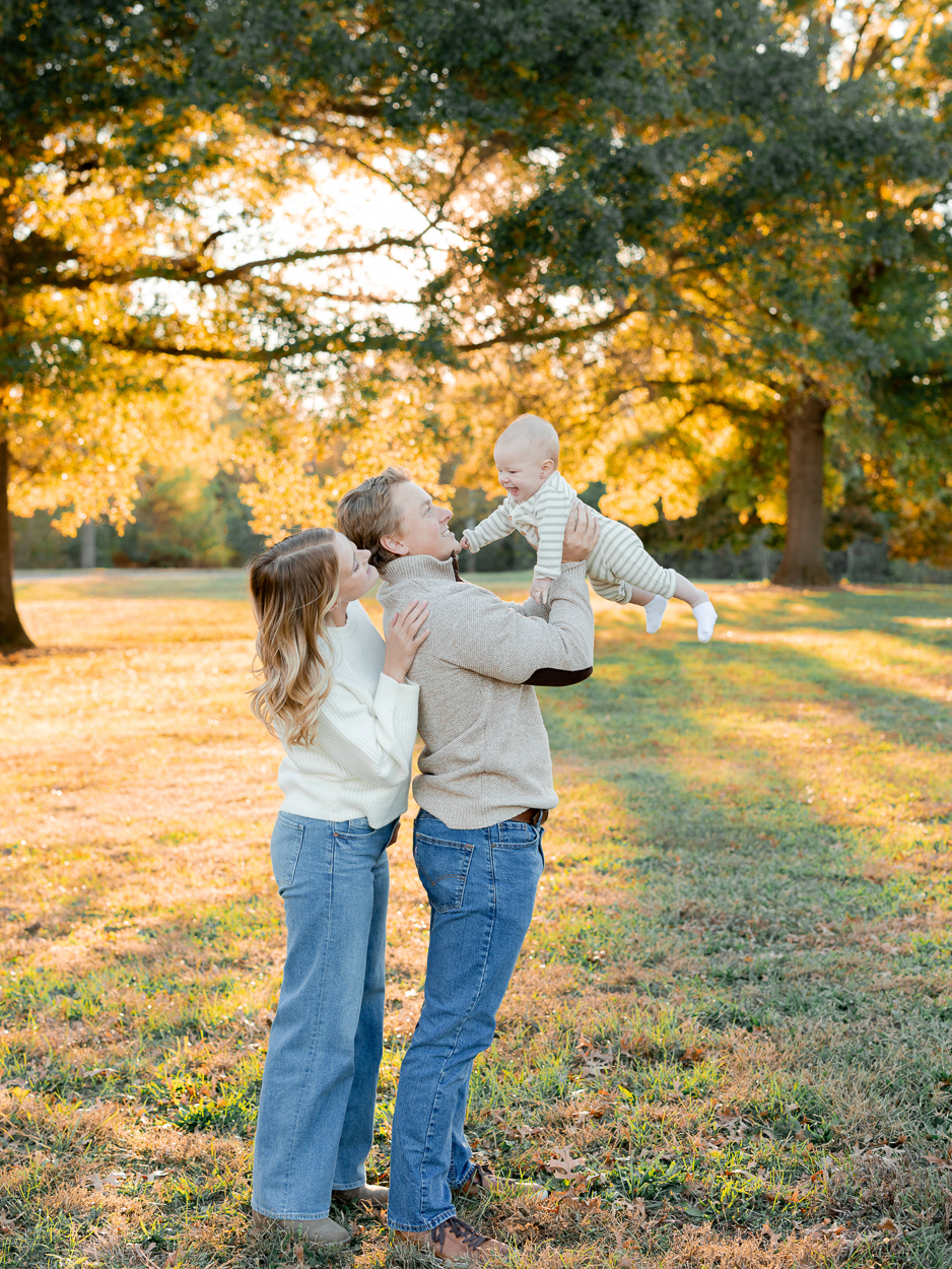 Parents standing under a large golden tree as they share a kiss while holding their baby, photographed by Love Tree Studios for golden sunset family photos in Columbia, MO.