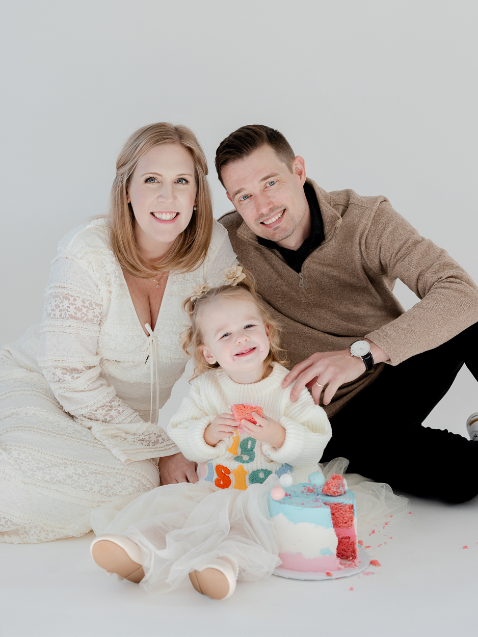 Family portrait with parents smiling beside toddler and pastel cake in Columbia Missouri studio.