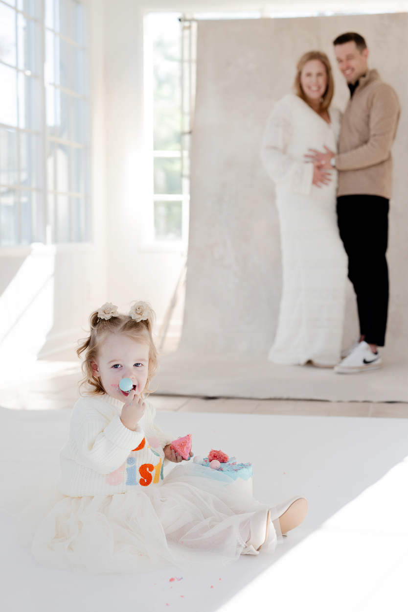 Toddler girl enjoying cake while parents embrace in background of Columbia Missouri natural light studio.