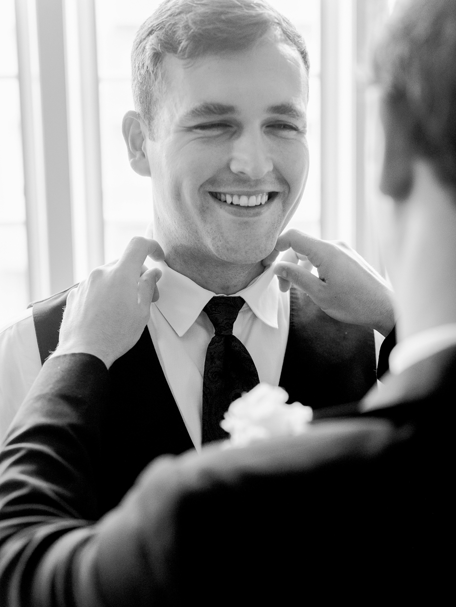 Groom smiling as his boutonniere and tie are straightened during getting ready moments at a Columbia Missouri church wedding.