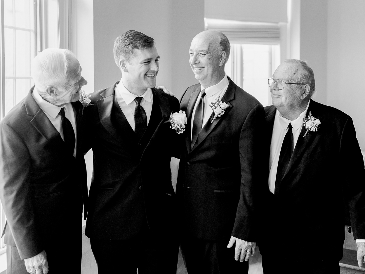 Groom standing with father and grandfathers in black suits sharing a laugh before their Columbia Missouri church wedding ceremony.