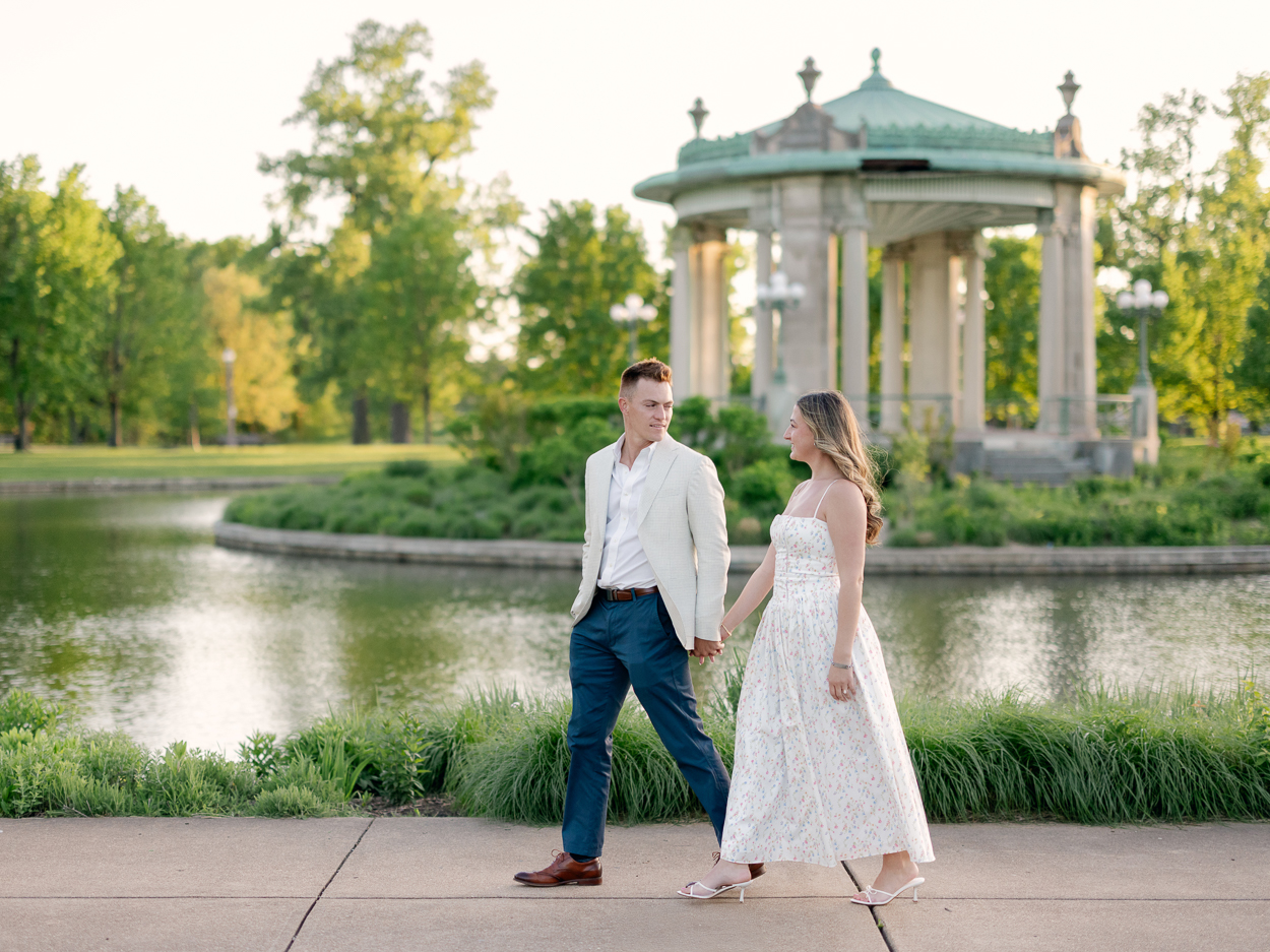 St. Louis engagement photographer captures couple walking hand in hand in front of Pagoda Lake and the Nathan Frank Bandstand at Pagoda Circle in Forest Park.