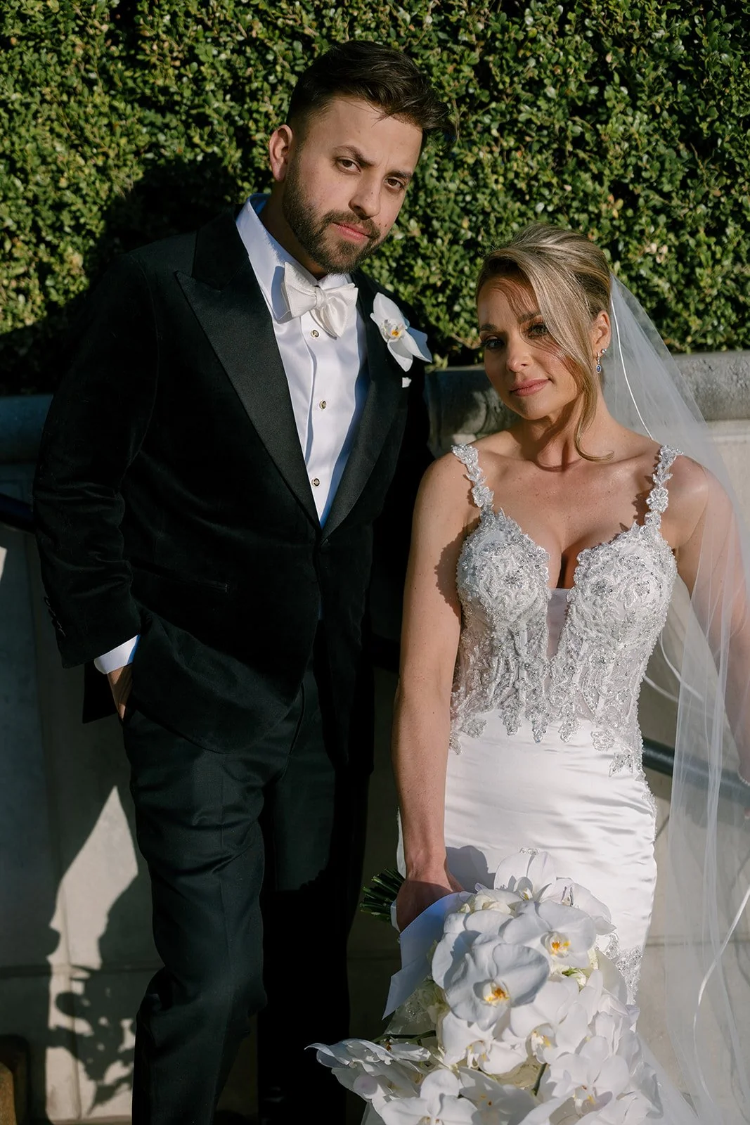 Bride and groom standing close together in front of a hedge wall, bride holding a white orchid bouquet, wedding photography by The Youngs.