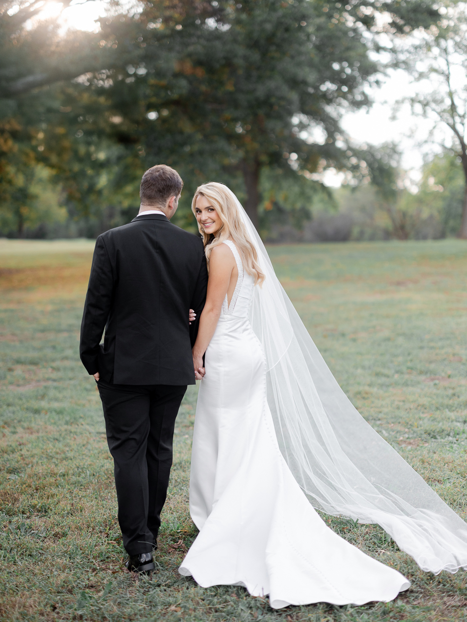 Bride smiling back over her shoulder while holding groom's arm, walking through an open field with a long veil trailing, St. Louis wedding photography by Love Tree Studios.