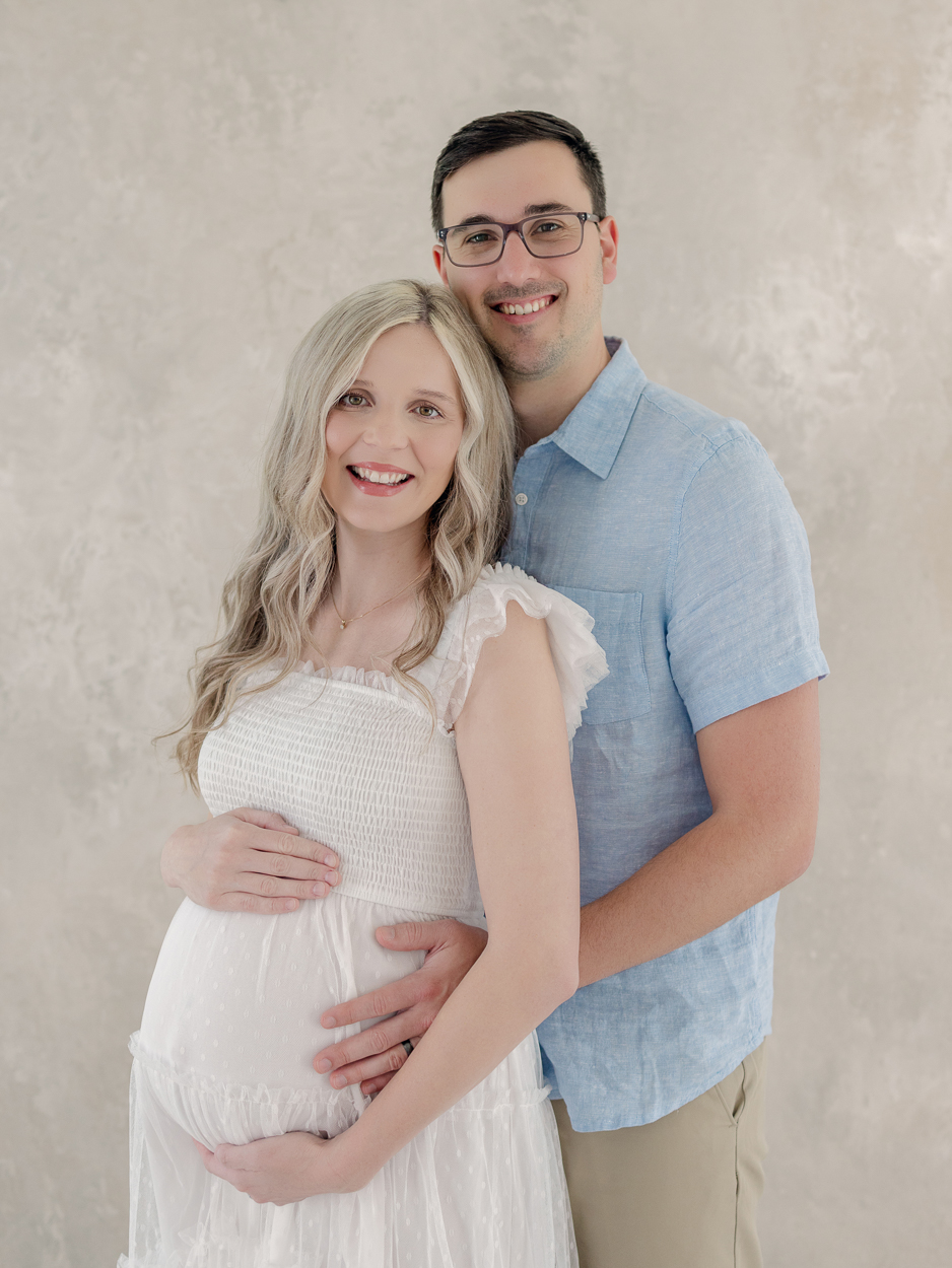 Expecting couple smiles at camera while husband stands behind pregnant wife cradling her baby bump during studio maternity session at Love Tree Studios Columbia MO.