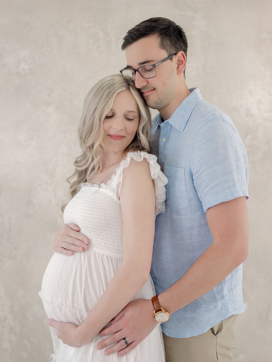 Expecting couple stands cheek to cheek looking down at baby bump during natural light maternity session at Love Tree Studios in Columbia Missouri.