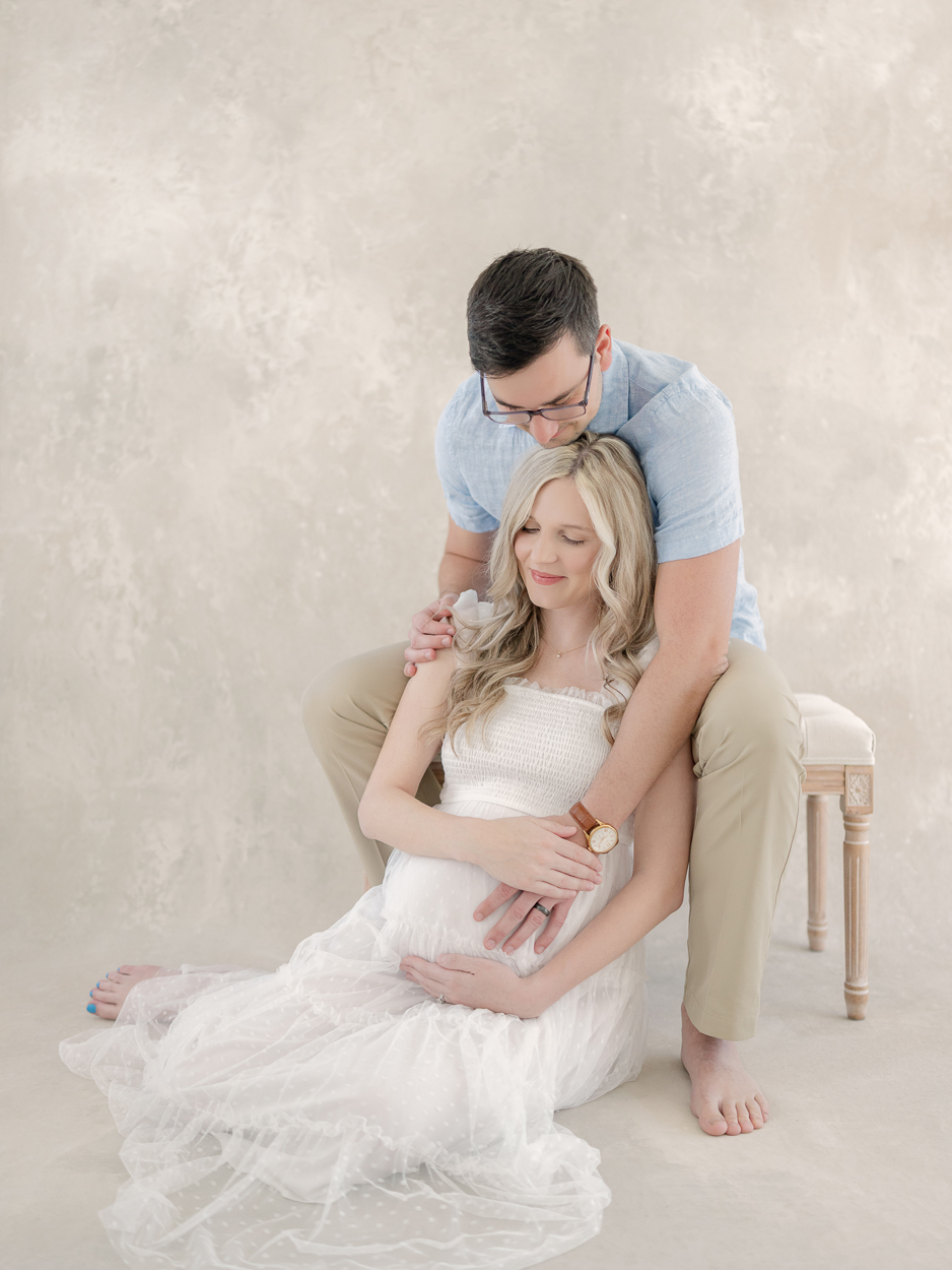 Expectant mother in white tulle dress sits on studio floor while husband kisses her head and wraps his arms around her during maternity session at Love Tree Studios in Columbia MO.