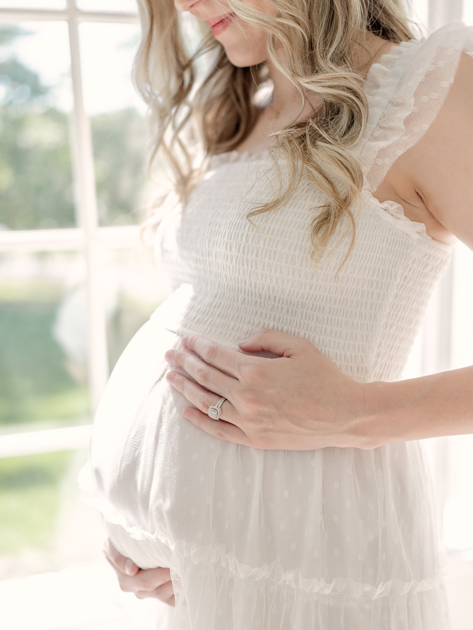 Close up of pregnant woman cradling baby bump in white smocked tulle dress with cushion cut diamond engagement ring visible during window maternity portraits at Love Tree Studios Columbia MO.