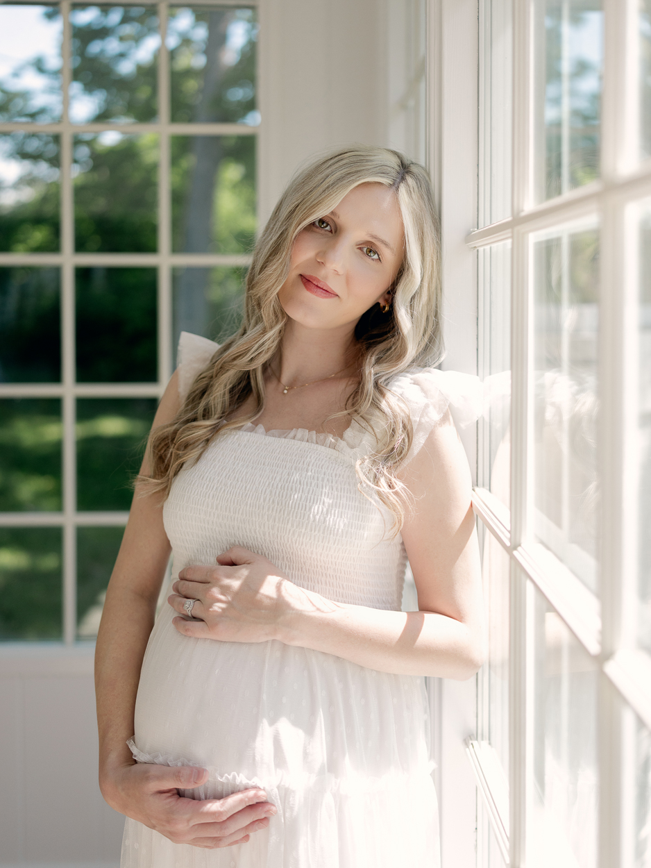 Pregnant woman in white smocked tulle dress leans against window frame looking at camera while cradling baby bump during maternity session at Love Tree Studios Columbia MO.