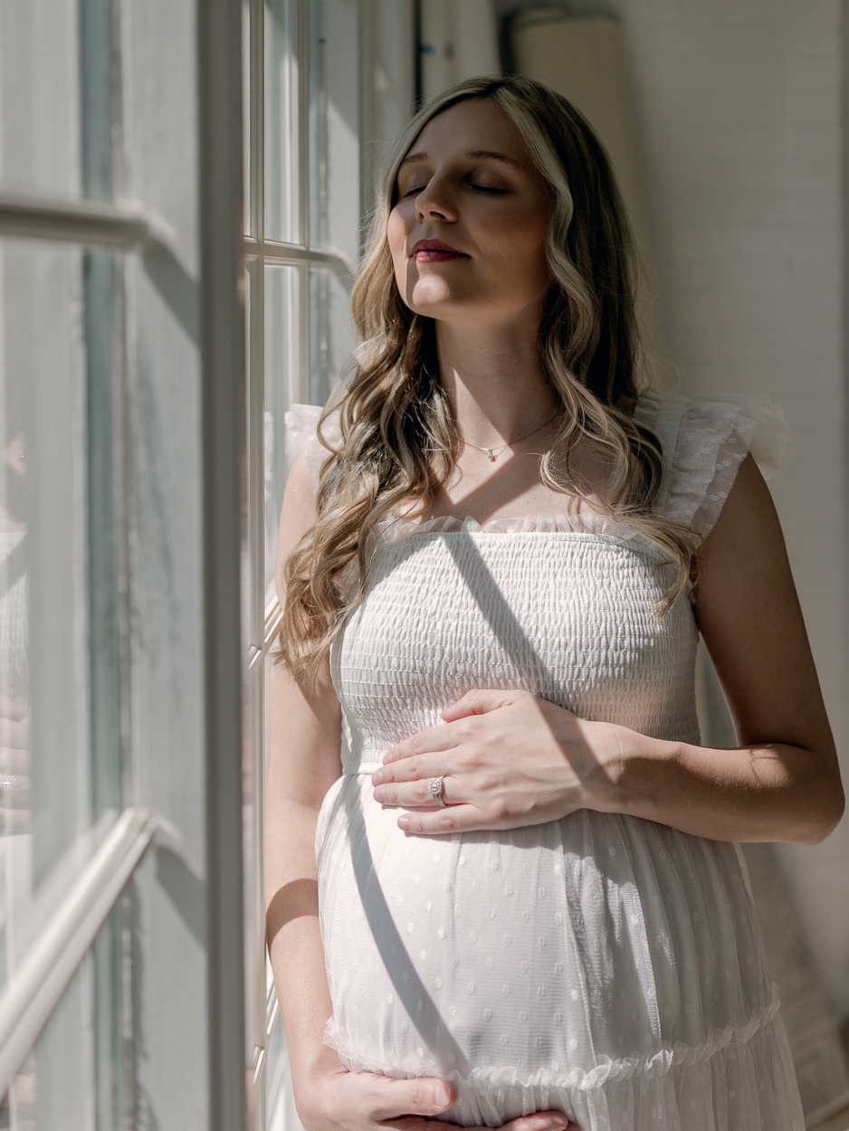 Pregnant woman in white tulle dress stands at window with eyes closed and sunlight across her face while cradling baby bump during maternity session at Love Tree Studios Columbia MO.