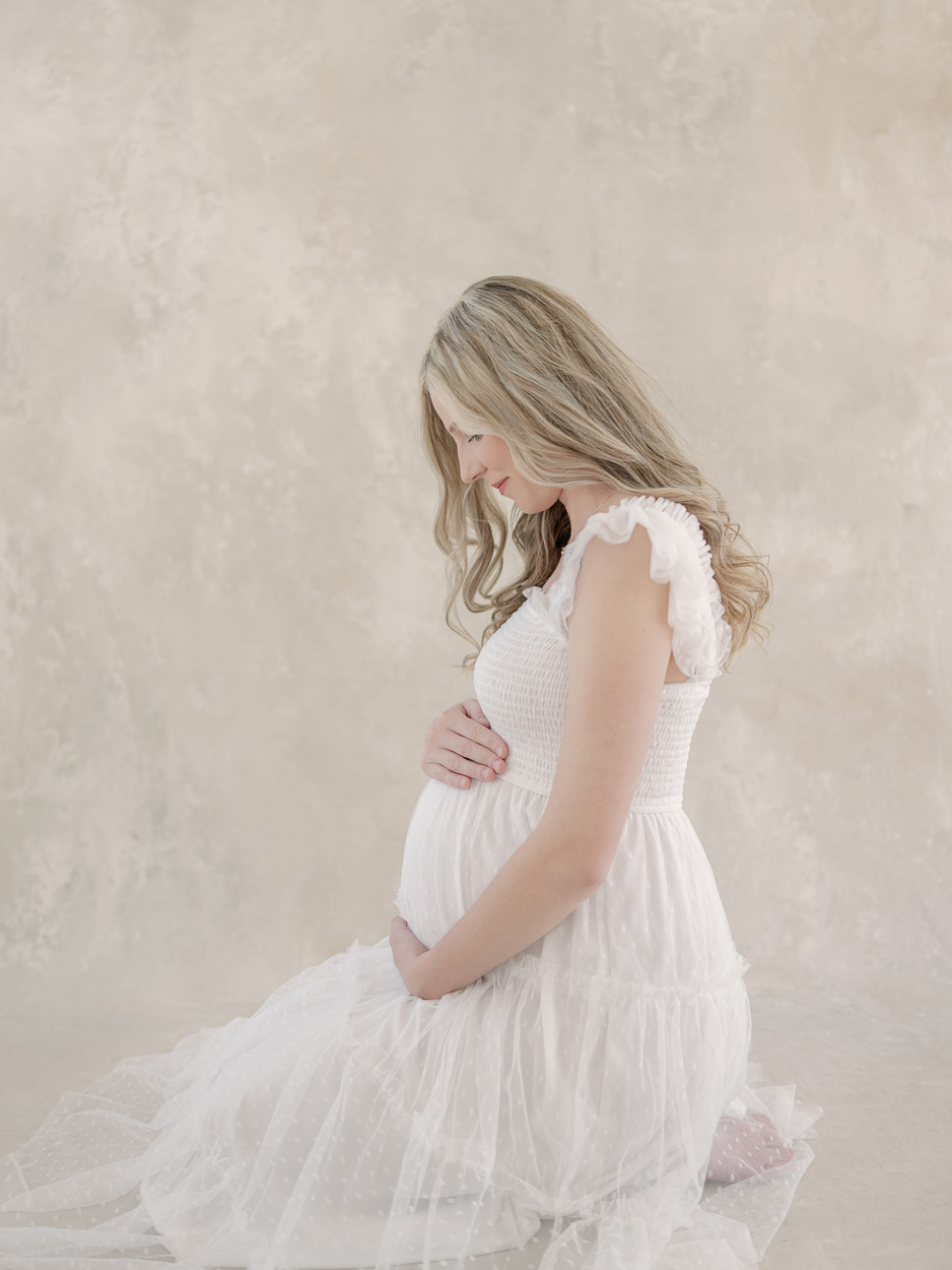 Pregnant woman in white tulle dress kneels on studio floor cradling her baby bump during maternity session at Love Tree Studios Columbia MO.
