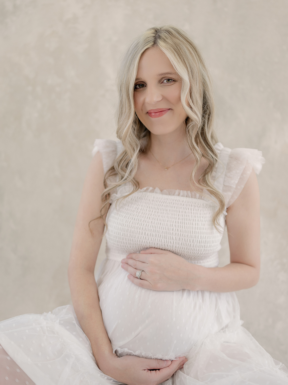 Pregnant woman in white smocked tulle dress sits on studio floor smiling at camera with diamond engagement ring visible during maternity session at Love Tree Studios Columbia MO.