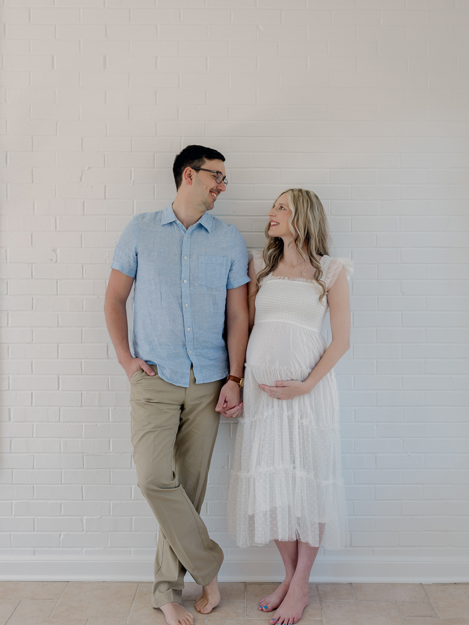 Expecting couple stands against white brick wall smiling at each other while holding hands during indoor maternity session at Love Tree Studios Columbia MO.