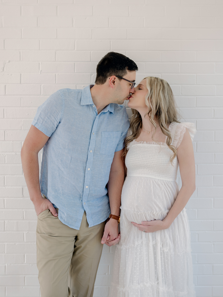 Expecting couple kisses while holding hands against white brick wall during natural light maternity session at Love Tree Studios in Columbia MO.