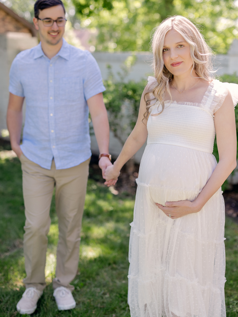 Pregnant woman smiles at camera while husband holds her hand and walks behind her during outdoor maternity portraits at Love Tree Studios Columbia MO.