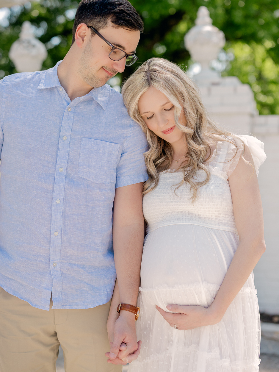 Expecting couple stands together with pregnant wife leaning on husband's shoulder looking down at baby bump in front of white stone balustrade during outdoor maternity session in Columbia MO.
