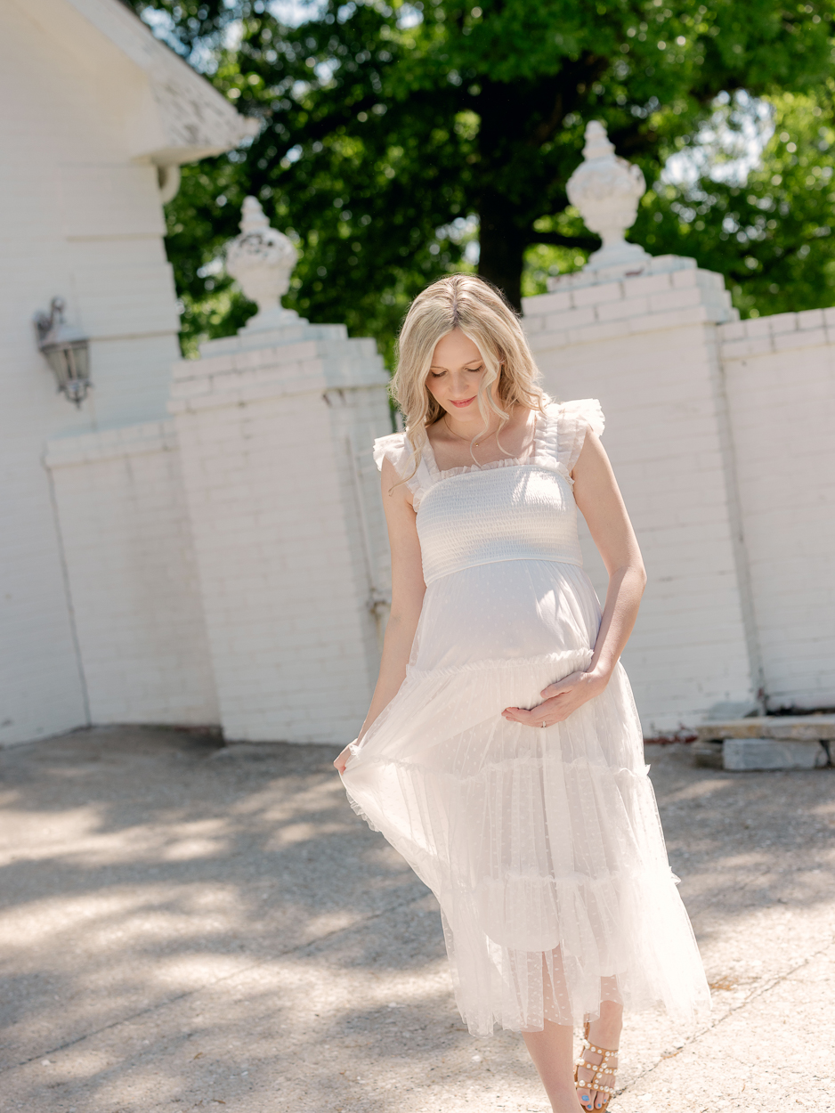 Pregnant woman in white tiered tulle dress walks along white brick wall looking down at baby bump during outdoor maternity session at Love Tree Studios in Columbia Missouri.