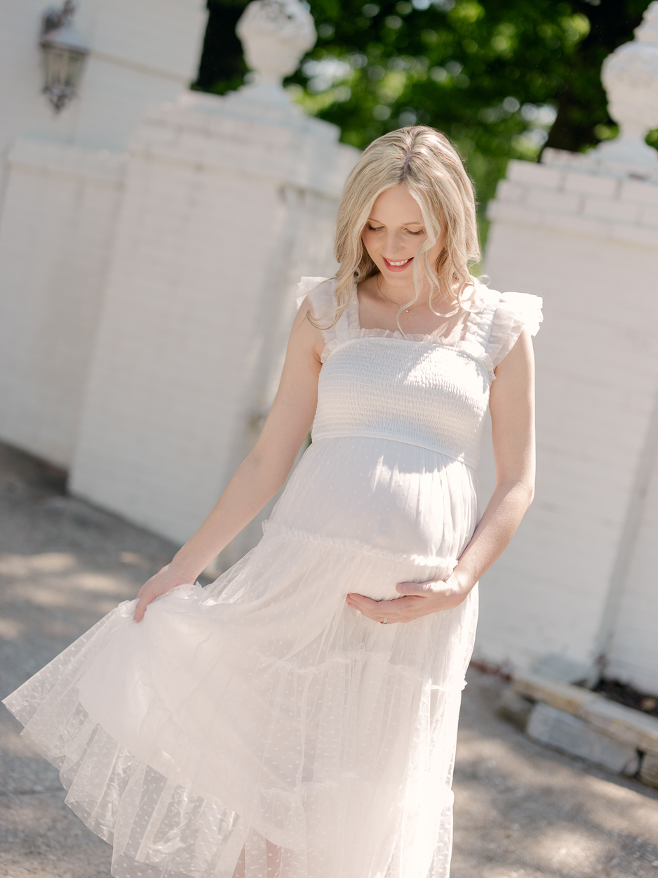 Pregnant woman in white tiered tulle dress smiles down while swishing skirt in front of white stone balustrade during outdoor maternity portraits at Love Tree Studios Columbia MO.