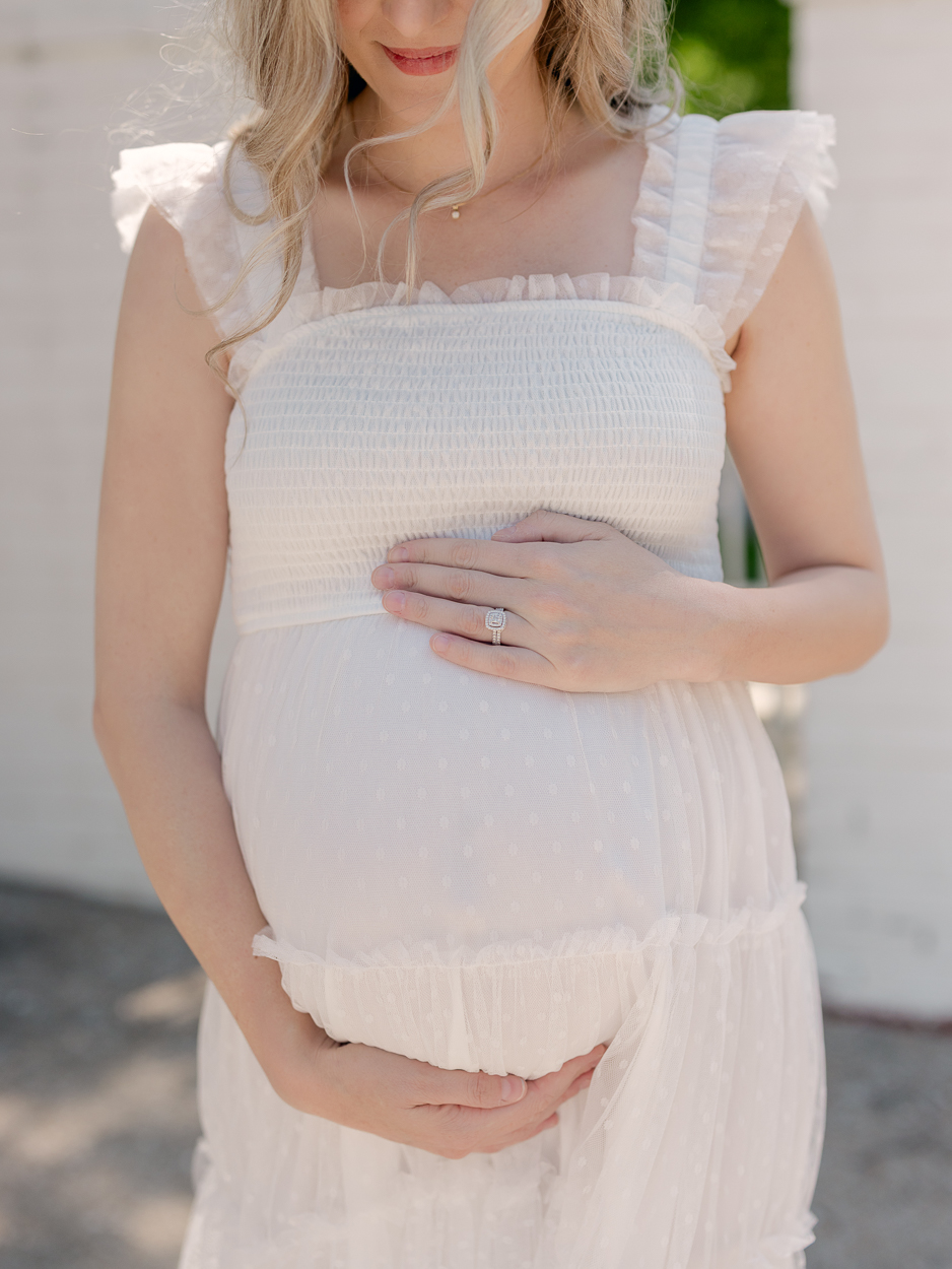 Close up of pregnant woman cradling baby bump in white dotted tulle dress with cushion cut diamond engagement ring visible during outdoor maternity session at Love Tree Studios Columbia MO.
