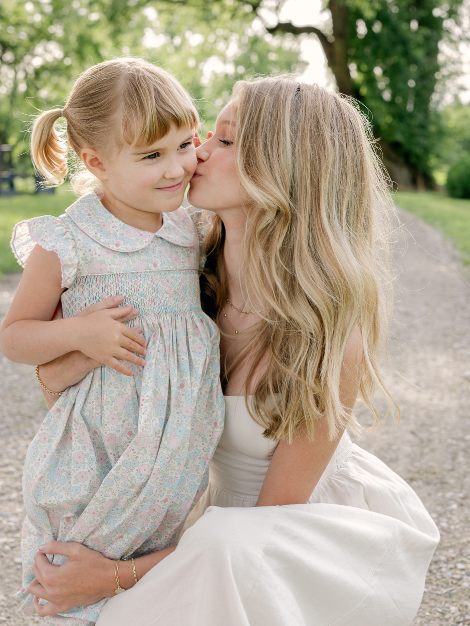 Teen girl kissing her little sister's cheek on a gravel path lined with trees.