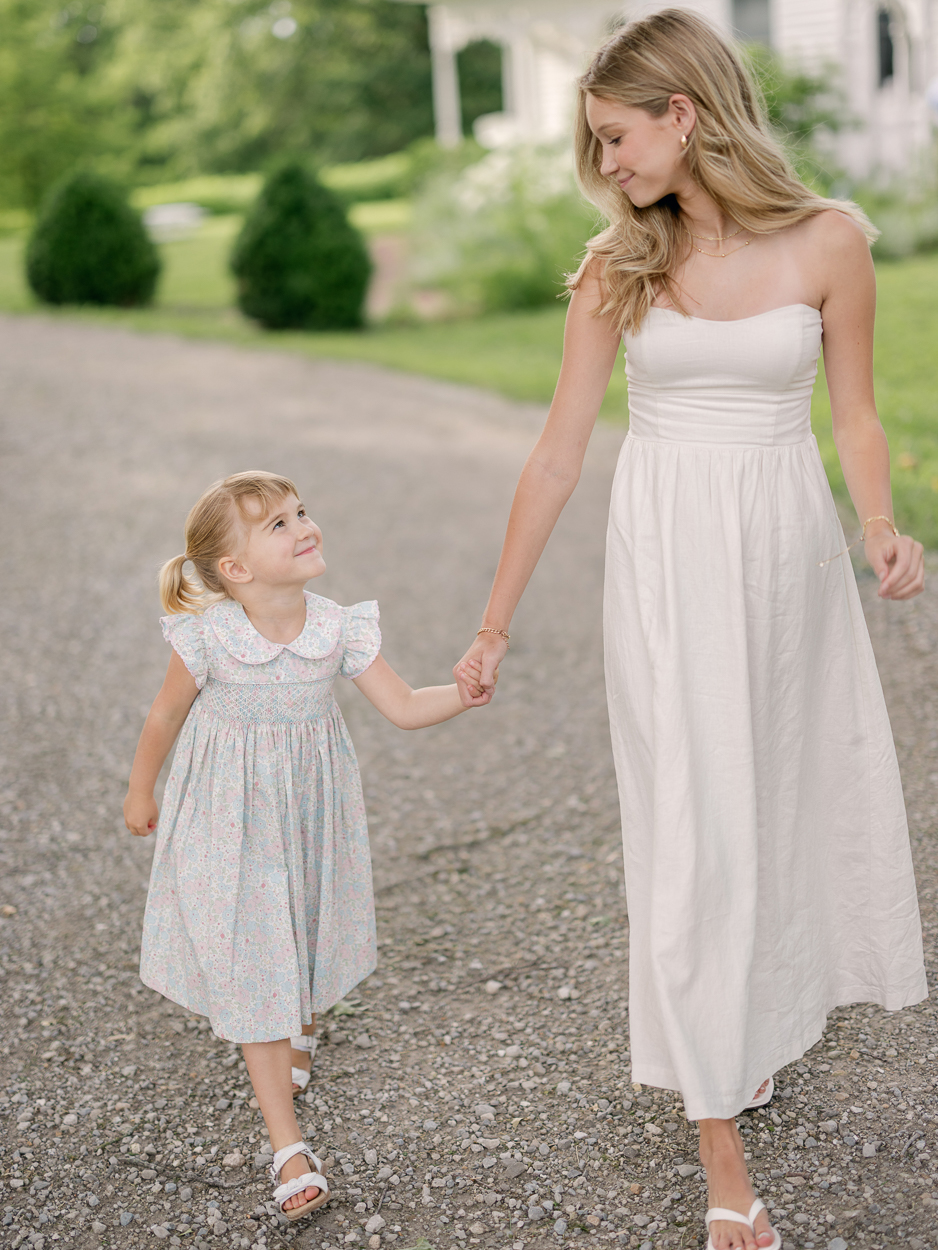 Little girl in floral smocked dress looking up at her older sister while holding hands, walking near farmhouse.
