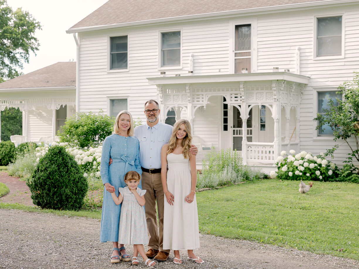 Family of four standing together in front of white Victorian farmhouse, Columbia MO family photographer.