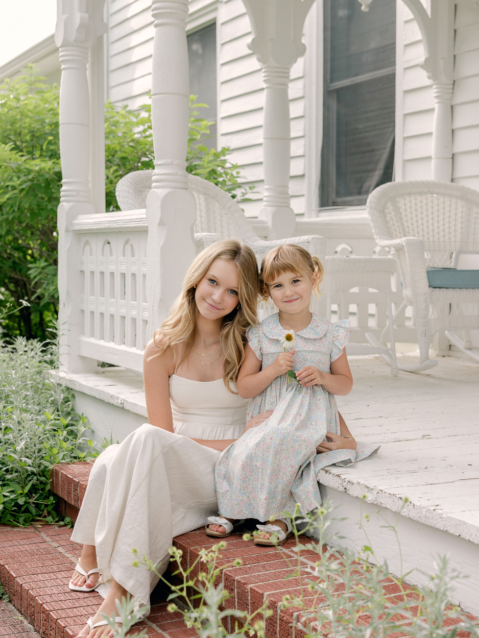 Two sisters sitting together on brick porch steps, younger sister holding a small white flower.