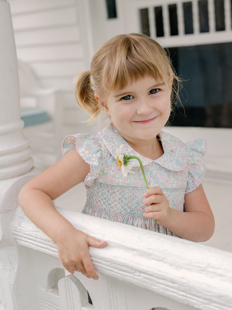 Little girl in floral dress holding a daisy, leaning on a white porch railing.