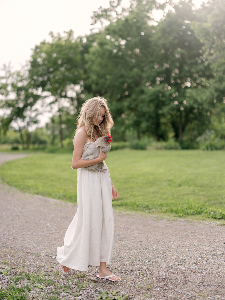 Teen girl in white dress walking outdoors while holding a pet chicken.