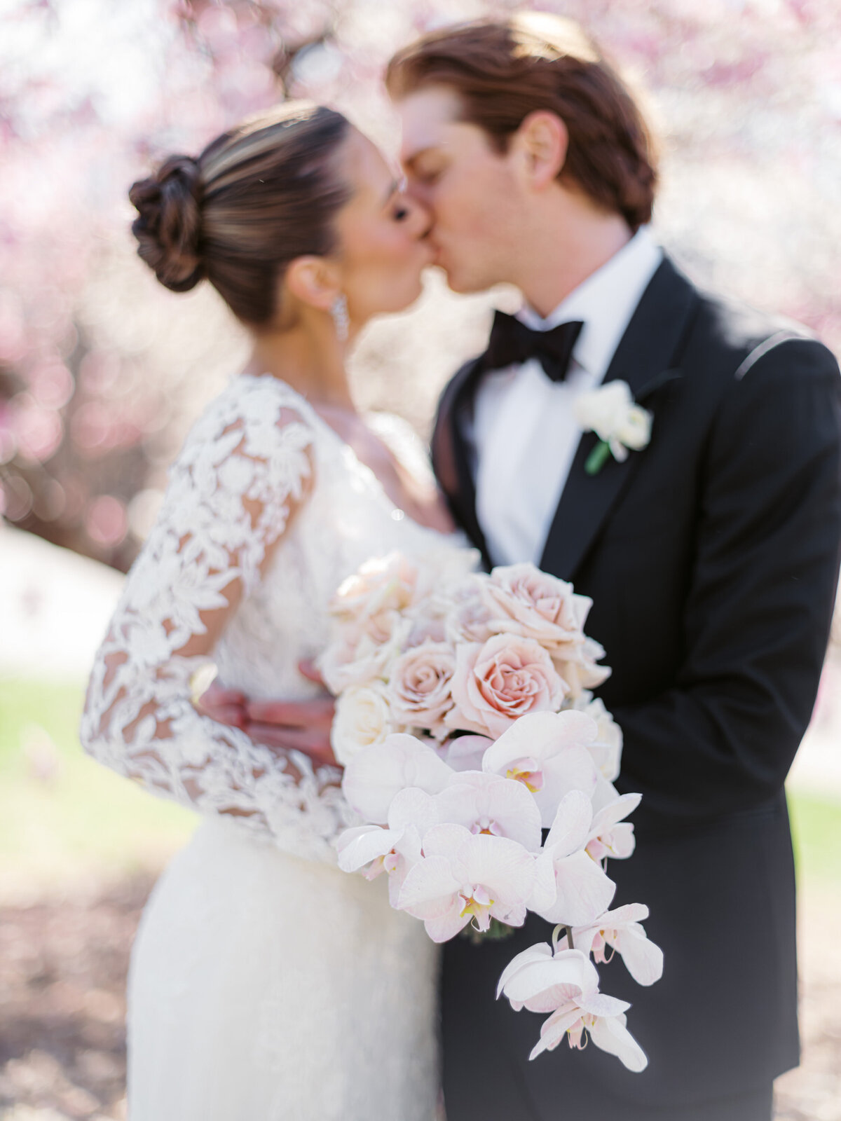 Close-up of bride and groom kissing, bride holding a bouquet of blush roses and orchids, cherry blossoms blurred in the background, wedding photography by Mike Cassimatis.