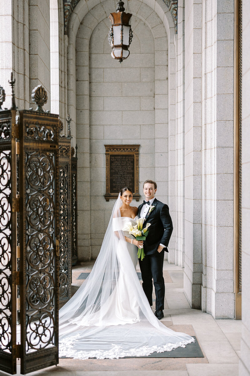 Bride and groom standing together beneath a stone archway at the St. Louis Central Library, bride holding a bouquet of calla lilies, wedding photography by Trent Broglin.